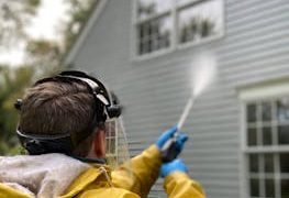 Person cleaning house siding with a pressure washer, wearing protective gear in a yellow raincoat.